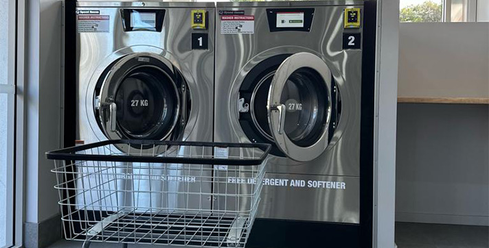 Two Extra large 27Kg stainless steel, cashless, touch screen operated, washing machines with an empty trolley and a bench