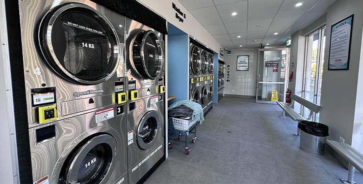 Coin laundry Merrylands with many washing machines and dryers, clean floor, trolley with clothes, card reader, bench, dustbin