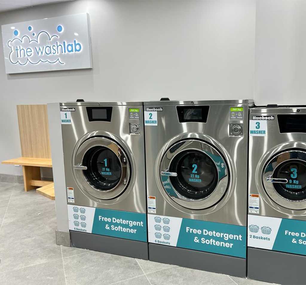 Three front-load self-service washers at a Wash Lab laundromat, offering free detergent and softener, with a bench and branded wall sign